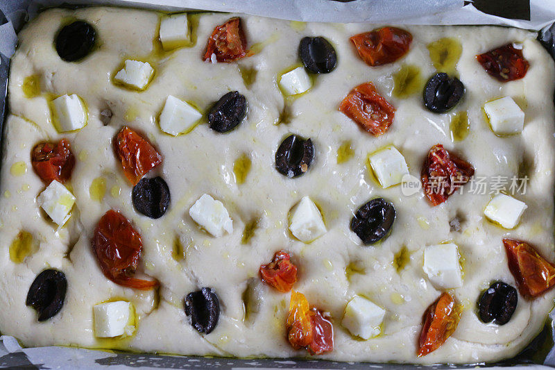 上图是一个自制的意大利佛卡夏面包，在烤盘里，内衬有防油纸/羊皮纸、绿橄榄、晒干的番茄和羊乳酪，淋上橄榄油，俯瞰风景